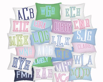 May include: A collection of rectangular throw pillows in various pastel and solid colours, each featuring a personalised monogram or name embroidered in contrasting thread. The pillows are arranged in a dense, overlapping cluster.