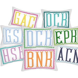 May include: A collection of eight rectangular white throw pillows, each featuring a scalloped border and a three-letter monogram in various colours including yellow, teal, light blue, green, lavender, orange, and dark navy blue.