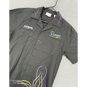 May include: A black short-sleeve button-down shirt featuring purple lightning bolt embroidery on the collar, "Grand Opening 2012" text on the right chest, "Ramone" in script on the left chest, and colorful pinstripe designs at the bottom hem.