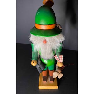 May include: A wooden nutcracker figurine with a green hat, white beard, and a red nose. The nutcracker is holding binoculars and a bundle of acorns.