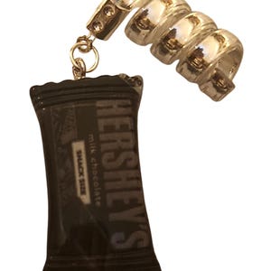May include: A silver charm bracelet with a Hershey's chocolate bar charm. The charm is shaped like a chocolate bar wrapper and has the Hershey's logo and the words "milk chocolate" and "snack size" printed on it.