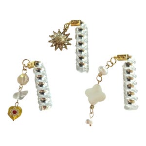 Puede incluir: Tres colgantes de plata y oro con diferentes encantos. El primer encanto es un girasol dorado, el segundo encanto es un corazón blanco con un centro rojo y el tercer encanto es un trébol de cuatro hojas blanco.