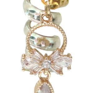 May include: A gold and silver ear cuff with a dangling charm featuring a crystal bow and a teardrop-shaped crystal.