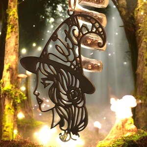 May include: A black metal silhouette of a witch with a pointed hat and flowing hair. The silhouette is hanging from a chain and is set against a blurry background of a forest.