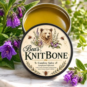 May include: A round metal tin of comfrey salve, labeled "Bea's KNITBONE." The lid features a bear illustration with bees and floral accents. The tin is open, revealing a yellow salve, surrounded by purple flowers.