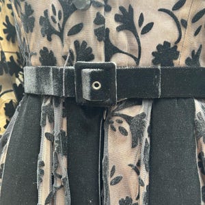 May include: Close-up of a black velvet belt with a square buckle and a black dress with floral patterns. The dress has a sheer overlay with black velvet floral designs and a button closure.