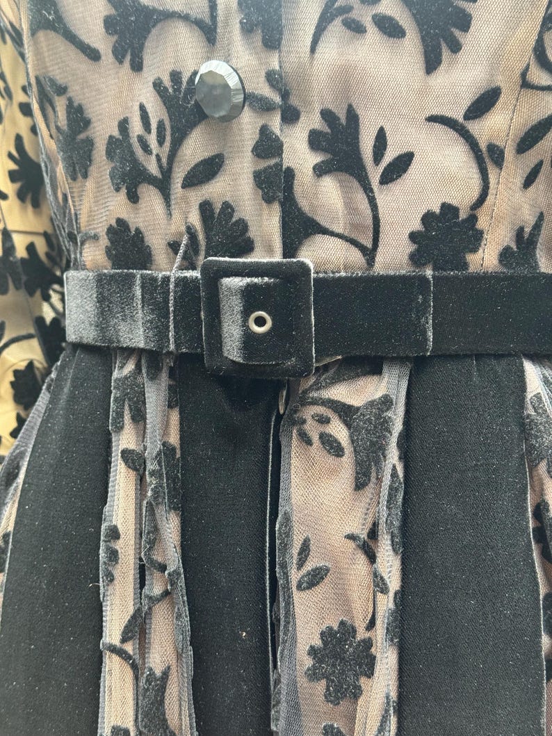 May include: Close-up of a black velvet belt with a square buckle and a black dress with floral patterns. The dress has a sheer overlay with black velvet floral designs and a button closure.