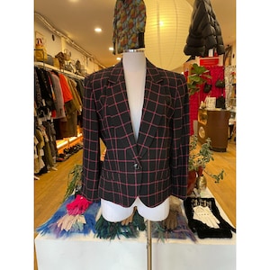 May include: Black blazer with a red check pattern, displayed on a mannequin. The blazer features a single-button closure and a classic notched collar. The background shows a vintage clothing store with racks of clothing and accessories.
