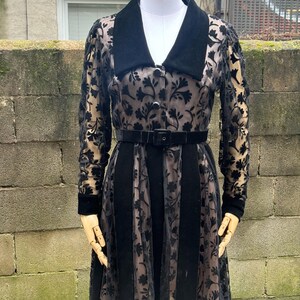 May include: A black vintage dress with a velvet collar and belt. The dress features a floral pattern on sheer sleeves and skirt panels. The dress is displayed on a mannequin.