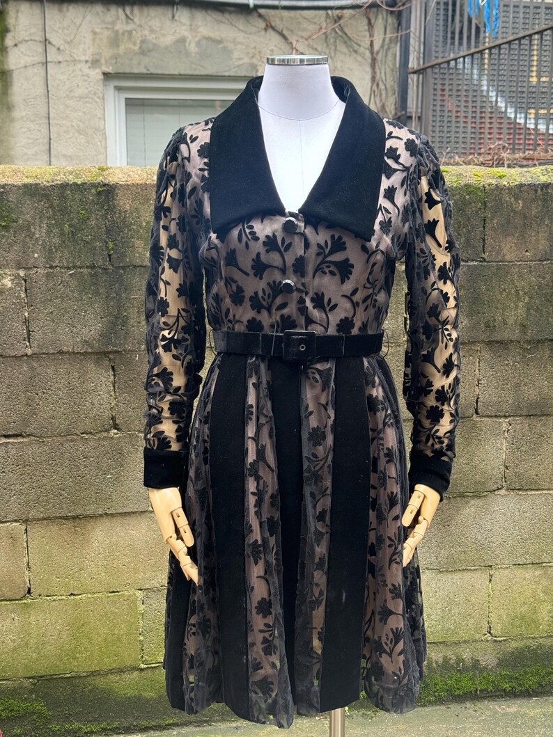 May include: A black vintage dress with a velvet collar and belt. The dress features a floral pattern on sheer sleeves and skirt panels. The dress is displayed on a mannequin.
