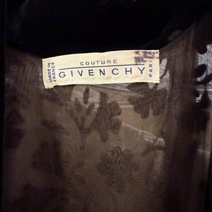 May include: Close-up of a black garment with a velvet collar. The label reads "COUTURE GIVENCHY PARIS" and "MADE IN FRANCE". The garment has a sheer, patterned layer underneath.
