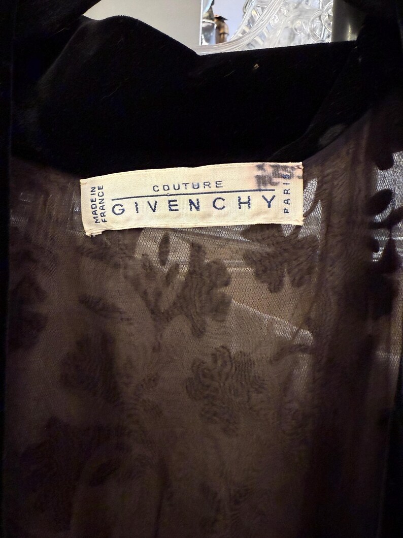 May include: Close-up of a black garment with a velvet collar. The label reads "COUTURE GIVENCHY PARIS" and "MADE IN FRANCE". The garment has a sheer, patterned layer underneath.