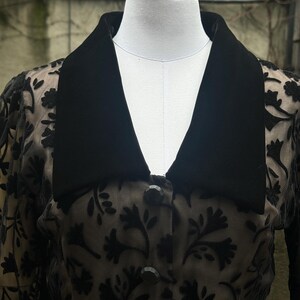 May include: A black, vintage-style jacket with a velvet collar and floral lace overlay. The jacket features a button-down front and a matching belt. The garment is displayed on a mannequin, showcasing its design and details.