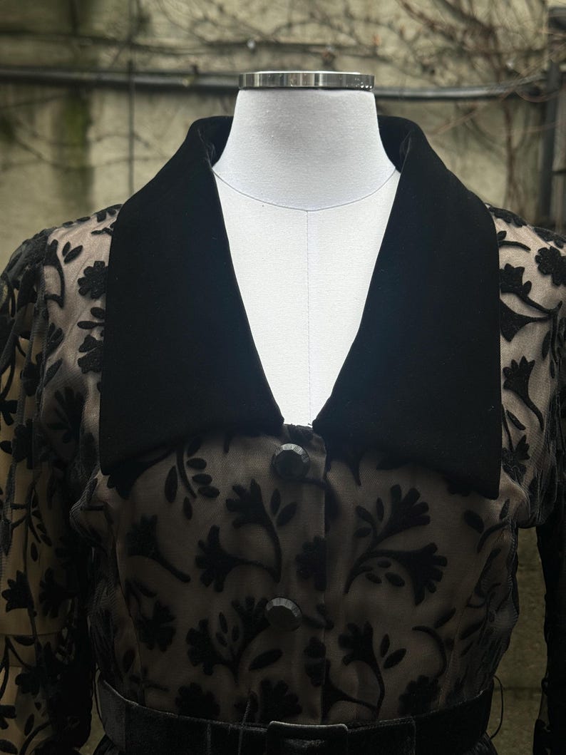 May include: A black, vintage-style jacket with a velvet collar and floral lace overlay. The jacket features a button-down front and a matching belt. The garment is displayed on a mannequin, showcasing its design and details.
