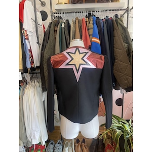 May include: Black jacket with a starburst design on the back. The starburst features a gold star outlined in pink, set against a burgundy and pink panel. The jacket is displayed on a mannequin in a clothing store.