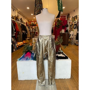 May include: Gold metallic pants with a drawstring waist and a relaxed fit. The pants are displayed on a mannequin in a boutique setting, showcasing a unique and eye-catching design. The pants have a shiny, crinkled texture.