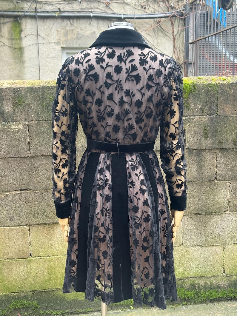 May include: A black vintage dress with a floral lace overlay. The dress features a velvet collar, a matching belt, and long sleeves. The skirt has vertical velvet panels alternating with the lace.