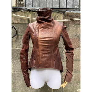 Puede incluir: Chaqueta de cuero marrón con cuello alto y mangas largas. La chaqueta tiene una silueta entallada y un diseño único, con un aspecto texturizado. Las mangas son de color marrón más oscuro. La chaqueta se exhibe en un maniquí.