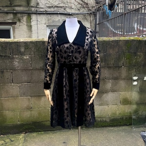 May include: Black velvet dress with floral lace sleeves and a matching collar. The dress features a belted waist and a knee-length skirt. The mannequin is displayed outdoors against a weathered brick wall.