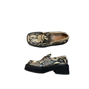 May include: Two pairs of shoes with a snakeskin pattern. One pair is a slip-on style, and the other has a chunky heel and platform sole. The shoes have a black and tan colour scheme.