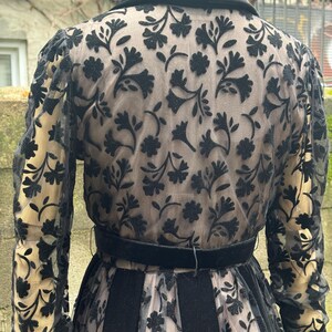 May include: A vintage black dress with a floral pattern overlay. The dress features a black velvet collar, a matching belt, and sheer sleeves. The floral design is visible on the sleeves and the back of the dress. The dress has a vintage aesthetic.