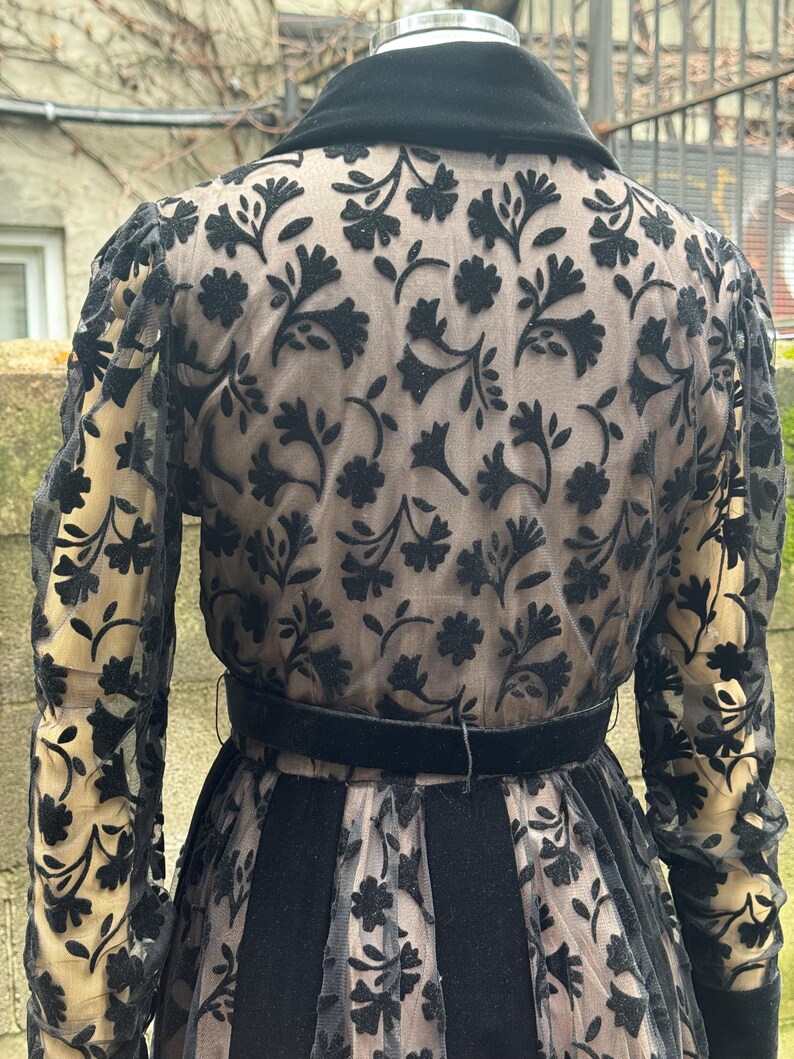 May include: A vintage black dress with a floral pattern overlay. The dress features a black velvet collar, a matching belt, and sheer sleeves. The floral design is visible on the sleeves and the back of the dress. The dress has a vintage aesthetic.