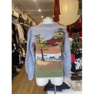 May include: A pale blue jumper featuring a landscape design. The design includes trees, a fence, and a mountain range in shades of green, brown, pink, and blue. The jumper has long sleeves and a crew neck.