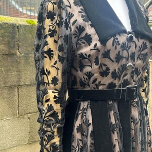 May include: A vintage black dress with a floral pattern overlay. The dress features a black velvet collar, belt, and cuffs. The sheer sleeves and skirt overlay reveal a beige underlay, creating a layered effect. The dress is buttoned down the front.
