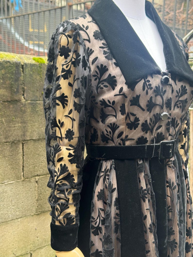 May include: A vintage black dress with a floral pattern overlay. The dress features a black velvet collar, belt, and cuffs. The sheer sleeves and skirt overlay reveal a beige underlay, creating a layered effect. The dress is buttoned down the front.