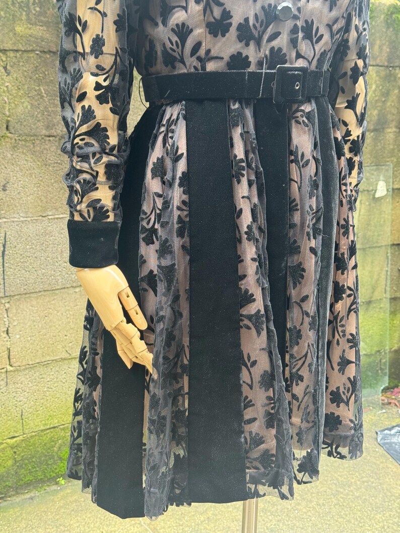 May include: A black dress with a velvet bodice and skirt panels, accented with sheer floral lace. The dress features long sleeves with velvet cuffs and a matching belt. The dress is displayed on a mannequin.