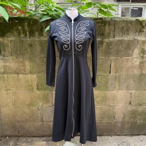 May include: Black long-sleeved dress with a full-length zip and a high collar. The dress has white embroidered wing-like designs on the chest. The dress is displayed on a mannequin against a brick wall background.