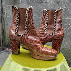 May include: Brown leather ankle boots with a snakeskin pattern. The boots feature a platform sole and a chunky heel. The top edge is trimmed with a leopard print. The boots are displayed on a green surface. The heels are approximately 10 cm high.