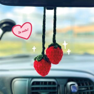 Puede incluir: Dos fresas de crochet rojas colgando de un espejo retrovisor en un coche. Las fresas tienen hojas verdes y están atadas a cuerdas verdes. Un adhesivo en forma de corazón rosa con el texto "So cute!" es visible en la imagen.