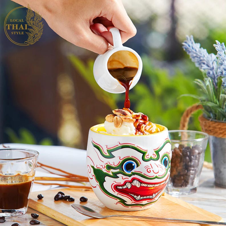 May include: A white ceramic mug with a colorful illustration of a mythical creature's face. The mug is filled with ice cream, peanuts, and caramel sauce. A hand is pouring espresso from a small white pitcher into the mug.