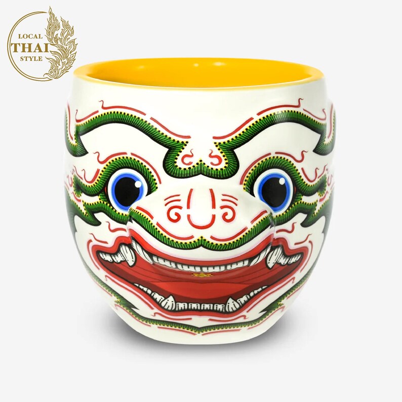 May include: White ceramic mug with a colorful illustration of a Thai mask. The mask has green, red, white, and blue details. The mug has a yellow interior.