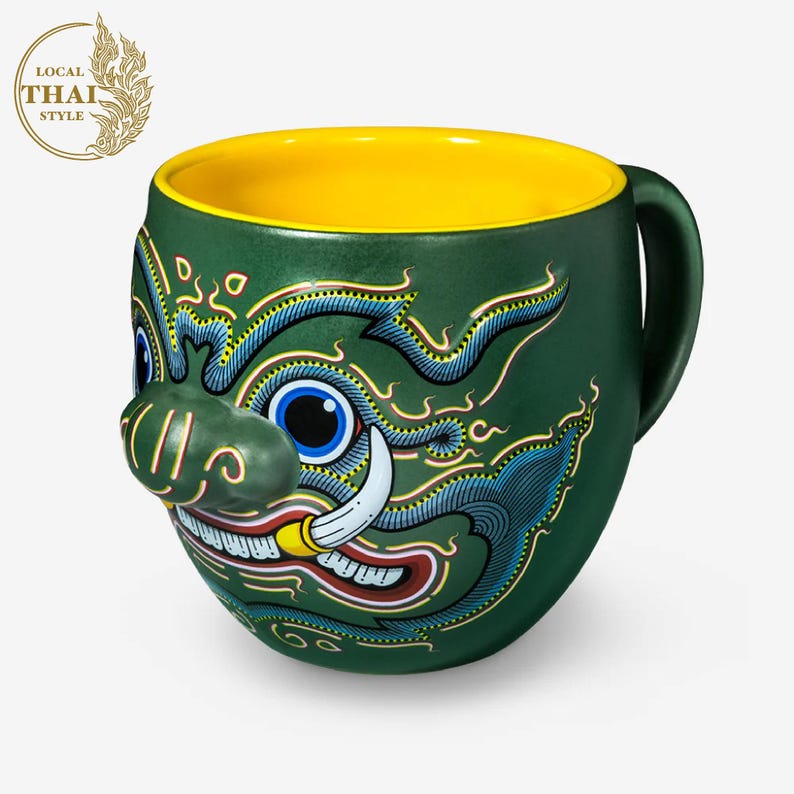May include: Green ceramic mug with a yellow interior and a colorful illustration of a mythical creature's face. The creature has a large, green nose, blue eyes, and a red mouth with sharp teeth. The mug has a handle on the right side.