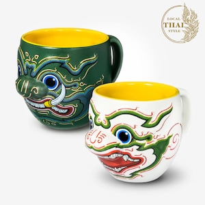 May include: Two ceramic mugs with a yellow interior and a colorful design of a Thai mythical creature's face. The mugs are green and white with red, blue, and yellow accents. The text "LOCAL THAI STYLE" is in a gold circle with a stylized leaf design.