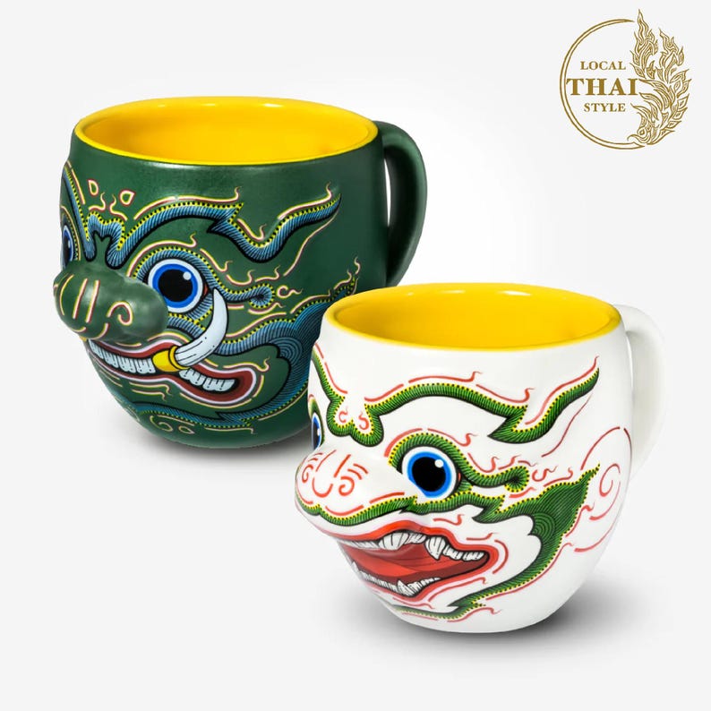 May include: Two ceramic mugs with a yellow interior and a colorful design of a Thai mythical creature's face. The mugs are green and white with red, blue, and yellow accents. The text "LOCAL THAI STYLE" is in a gold circle with a stylized leaf design.