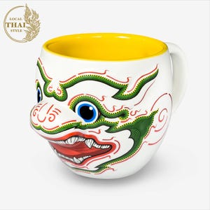 May include: White ceramic mug with a yellow interior and a colorful illustration of a Thai mythical creature with green, red, and white details. The creature has a large mouth with sharp teeth and a single blue eye.