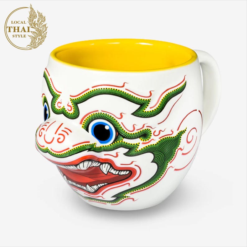 May include: White ceramic mug with a yellow interior and a colorful illustration of a Thai mythical creature with green, red, and white details. The creature has a large mouth with sharp teeth and a single blue eye.