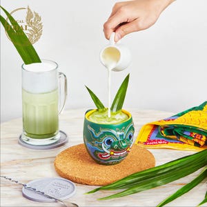 May include: A green ceramic mug with a colorful mythical creature design is being filled with a white liquid. The mug is sitting on a cork coaster with green leaves. A glass of green liquid with white foam is in the background.