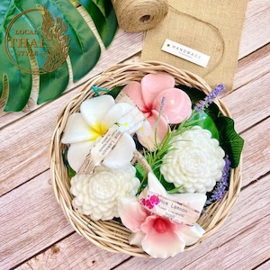 Peut inclure: Un panier en osier rempli de savons en forme de fleurs faits à la main. Les savons sont de différentes couleurs, notamment le blanc, le rose et le rose clair. Les savons sont étiquetés "Hom Lamoon" et "Plumeria".