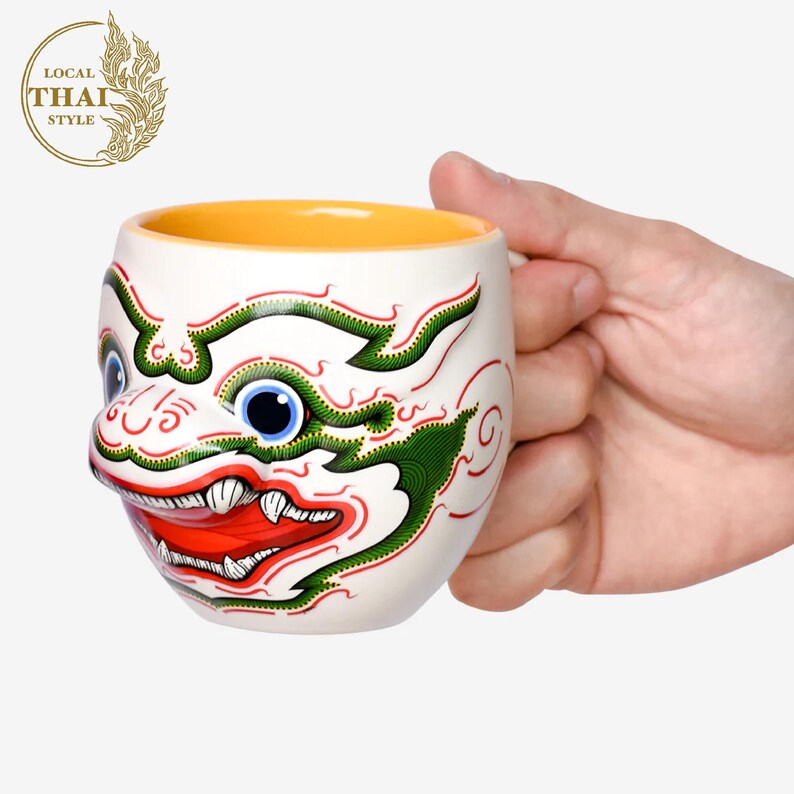 May include: White ceramic mug with a colorful illustration of a Thai mythical creature's face. The mug has a yellow interior and a handle.
