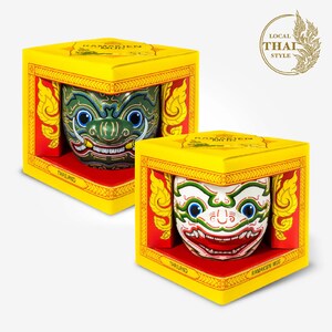 May include: Two yellow boxes with red and gold trim, each containing a ceramic mug with a colorful Ramakien mask design. The boxes have the text "Ramakien Mug" and "Thailand" printed on them. The boxes are on a white background.