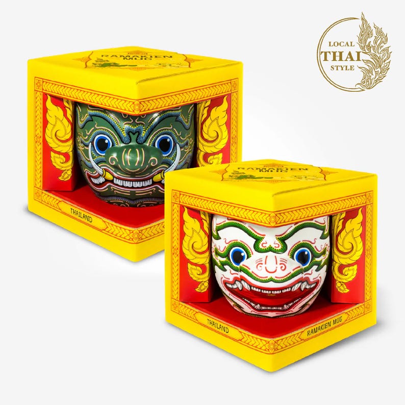 May include: Two yellow boxes with red and gold trim, each containing a ceramic mug with a colorful Ramakien mask design. The boxes have the text "Ramakien Mug" and "Thailand" printed on them. The boxes are on a white background.