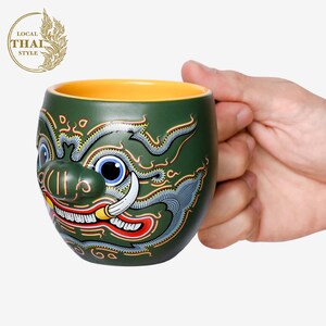 May include: Green ceramic mug with a colorful illustration of a mythical creature's face. The mug has a yellow rim and is being held by a hand.