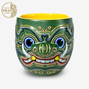 May include: Green ceramic mug with a yellow rim, featuring a detailed hand-painted design of a mythical creature with fangs, horns, and a fierce expression.