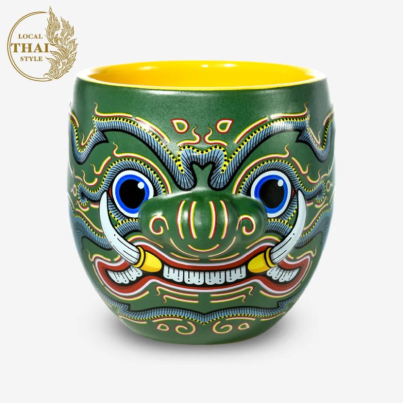 May include: Green ceramic mug with a yellow rim, featuring a detailed hand-painted design of a mythical creature with fangs, horns, and a fierce expression.