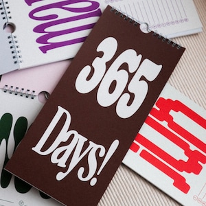 Può includere: Una collezione di calendari a spirale con testo in grassetto. Il calendario centrale è marrone con le parole "365 Days!" in bianco. Altri calendari presentano testo in viola, verde e rosso. Ogni calendario ha un foro per appenderlo.
