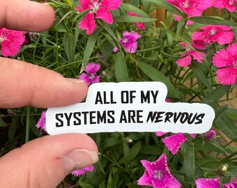May include: A white sticker with the text "ALL OF MY SYSTEMS ARE NERVOUS" in black, held in front of vibrant pink flowers and green foliage. The sticker is rectangular with rounded corners. The background includes a wooden fence.